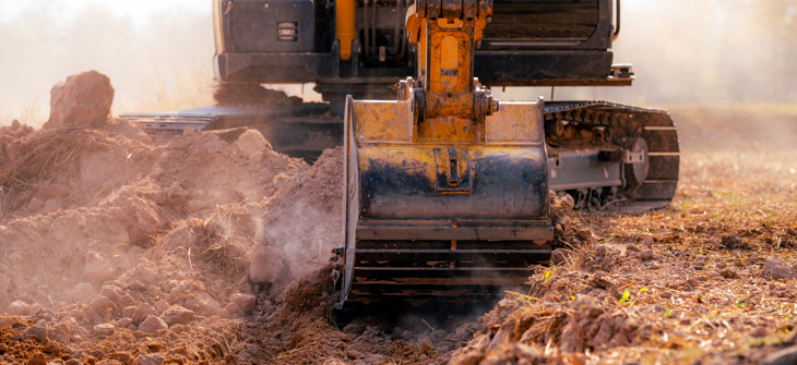 Close-up of an excavator digging into dry soil on a construction site, with dust rising around the machine.