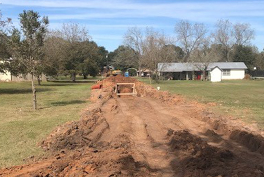 Trench dug in a grassy field with construction equipment in the background, flanked by trees and a few small buildings.