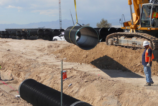 Workers install large drainage pipes using heavy machinery at a construction site with piles of pipes in the background.