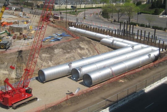 Large metal pipes are laid out at a construction site, with a red crane and equipment visible in the background.