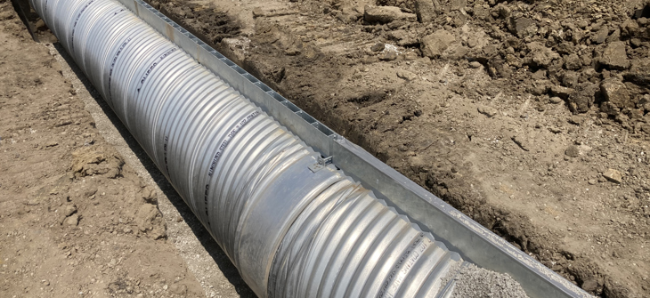 Large corrugated metal drainage pipe being installed in a trench surrounded by dirt at a construction site.