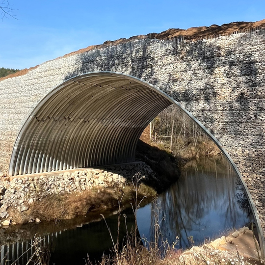 A wide stone bridge with an arched metal underpass spans a small river in a wooded landscape.