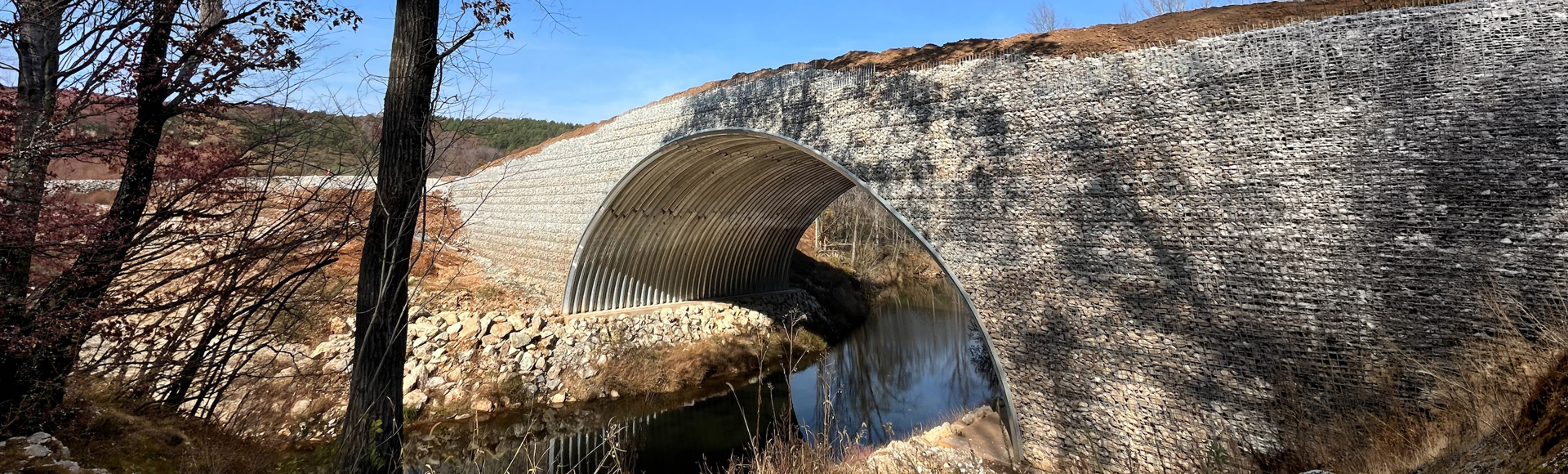 A wide stone bridge with an arched metal underpass spans a small river in a wooded landscape.