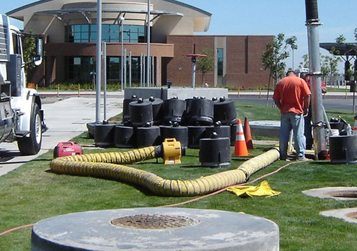Two workers perform maintenance near open manholes with safety cones, equipment, and a vacuum truck on a grassy area.