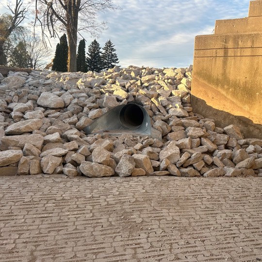 Concrete drainage pipe surrounded by rocks, located at the base of a sloped structure with stairs. Trees visible in the background.