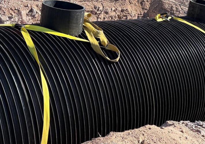 Large black ribbed tank secured with yellow straps is partially buried in a dirt trench at a construction site.
