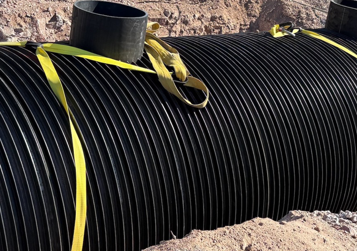 Large black ribbed tank secured with yellow straps is partially buried in a dirt trench at a construction site.