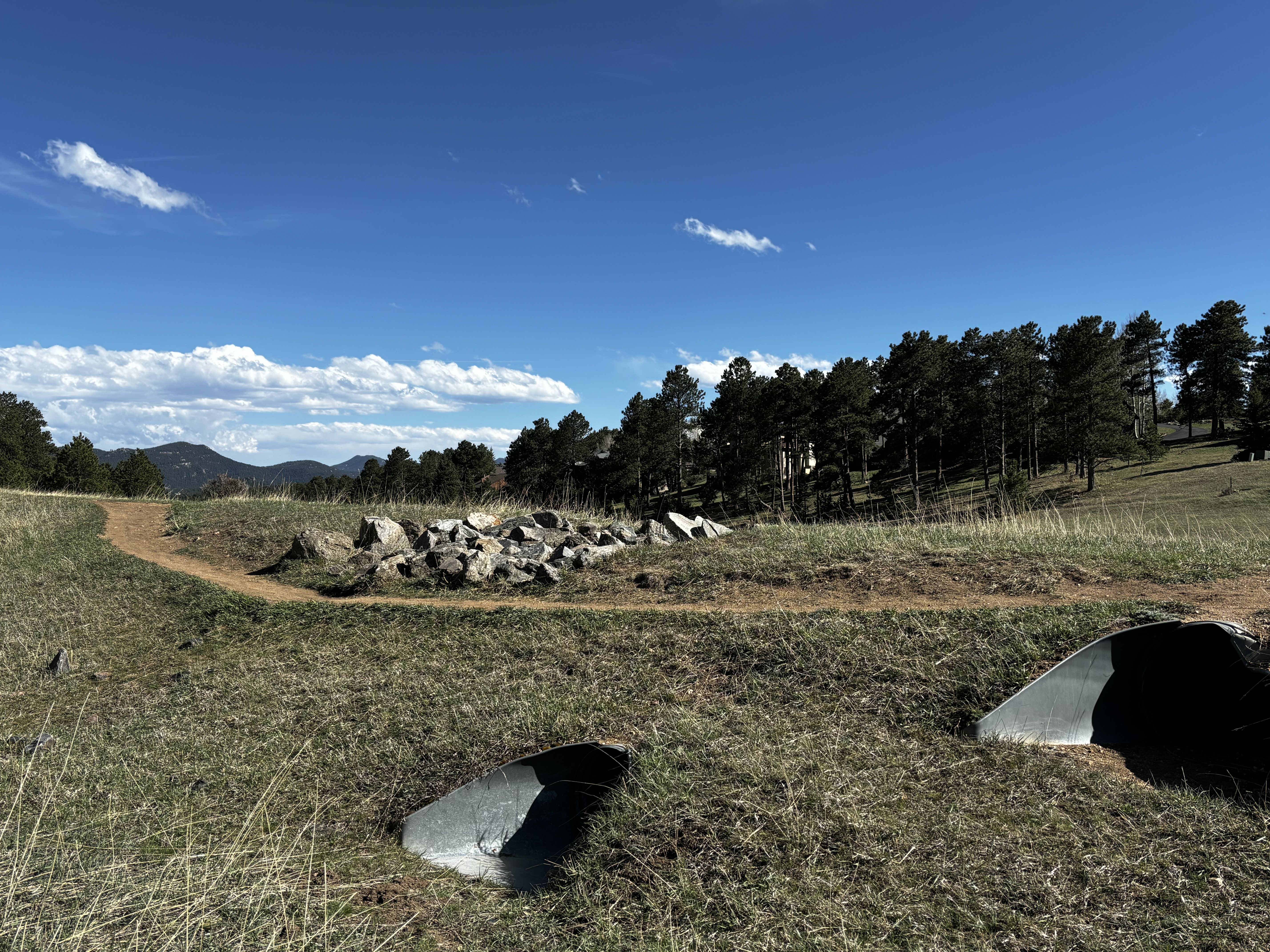 A dirt trail curves through grassy terrain with scattered rocks, pine trees, and metal culverts under a blue sky.
