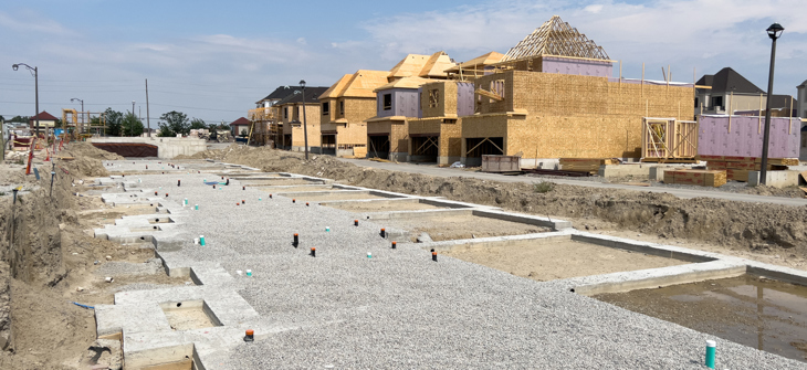 Row of new houses under construction with foundations and framing visible on a clear, sunny day.