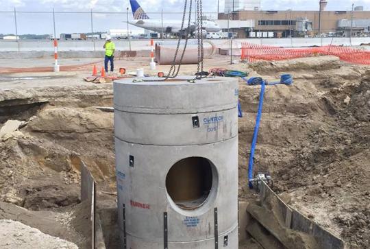 Construction workers are installing a large concrete drainage structure at an airport worksite with an airplane in the background.