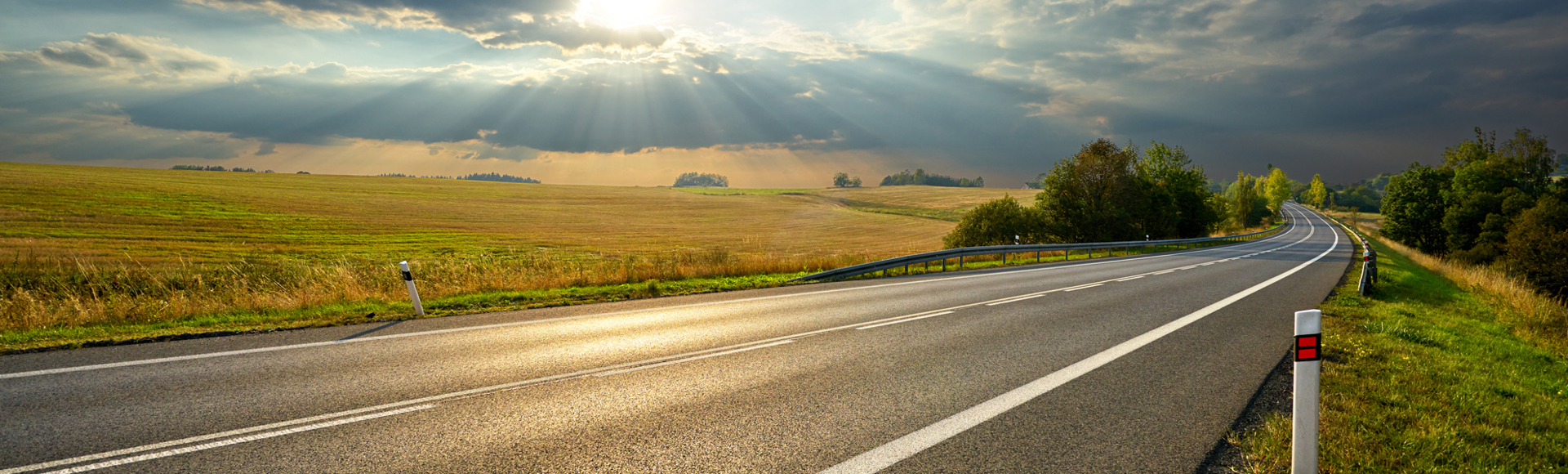 A paved rural road curves through open fields under a partly cloudy sky with sunlight streaming through the clouds.