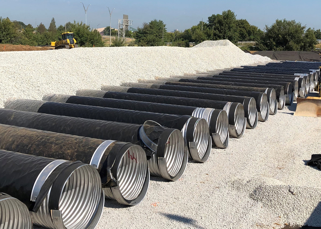 Several large corrugated metal pipes are lined up on a gravel surface at a construction site with earthmoving equipment visible.