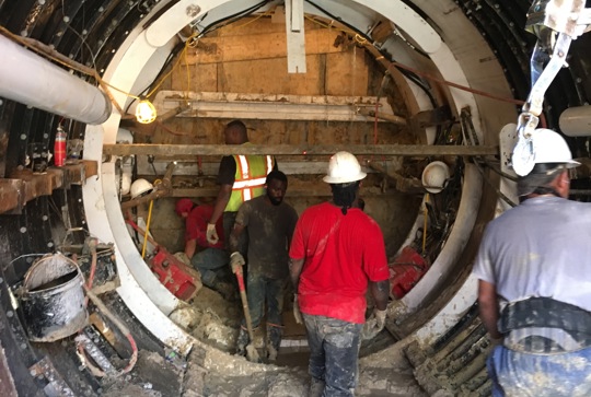 Workers in hard hats and safety gear performing construction inside a large tunnel with muddy ground and scaffolding.