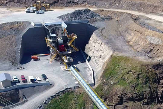 Aerial view of an industrial mining site with machinery, a conveyor belt, parked vehicles, and surrounding excavated earth.