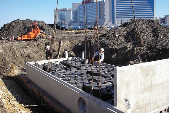 Construction workers install a large concrete structure with pipes at a construction site, with buildings in the background.
