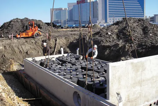 Construction workers install a large concrete structure with pipes at a construction site, with buildings in the background.