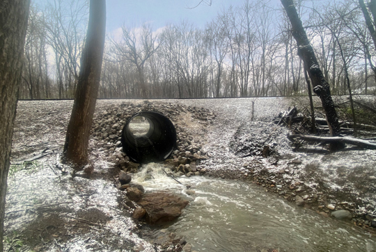 A culvert with water flowing through in a snowy forested area, with bare trees and a partially snow-covered ground.