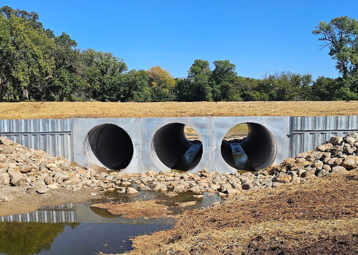 Three large concrete culverts channel water under a road, surrounded by rocks and vegetation on a clear day.