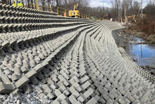 Concrete erosion control blocks are arranged on a sloped embankment beside a small body of water, with construction equipment nearby.
