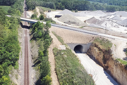 Aerial view of a construction site with a road bridge, a railway line, and a tunnel surrounded by trees and gravel piles.