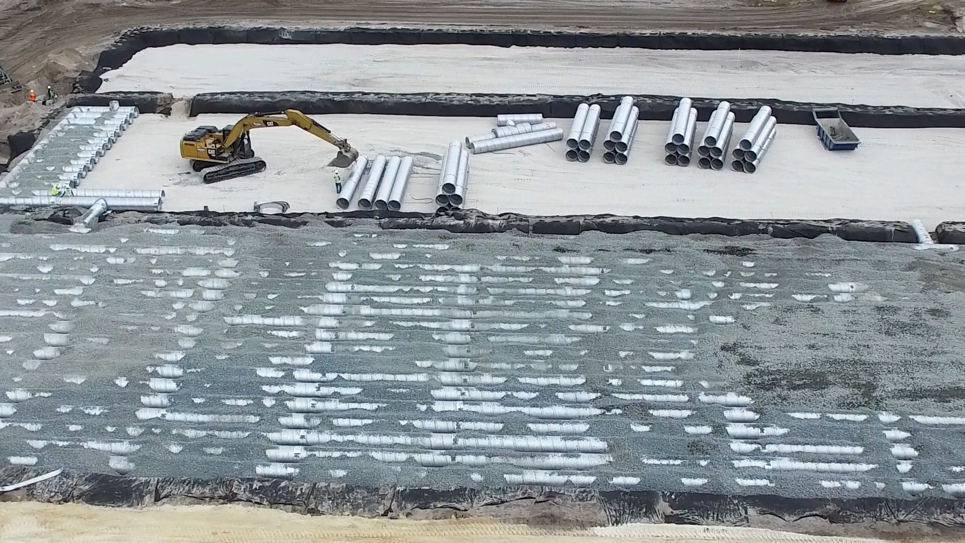 A construction site with an excavator and stacked pipes on gravel and sand layers, viewed from above.