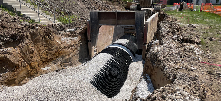 An excavator places gravel around a large drainage pipe in a trench at a construction site near a building and stairs.