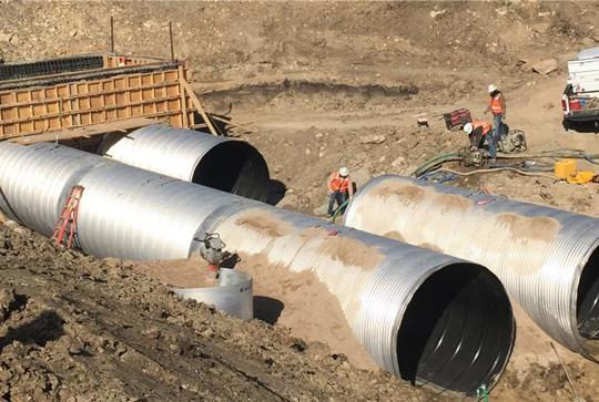 Construction site with three large metal drainage pipes, workers in orange vests, a white truck, and surrounding dirt terrain.