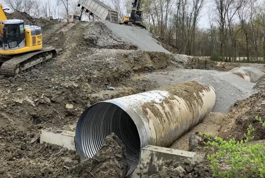 A construction site with a large metal drainage pipe being installed in a dirt embankment, surrounded by construction equipment.