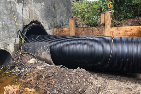 Corrugated black pipe being installed into a concrete culvert at a construction site, surrounded by dirt and wooden supports.