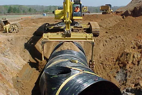Excavator placing a large black drainage pipe in a trench at a construction site, with additional machinery in the background.