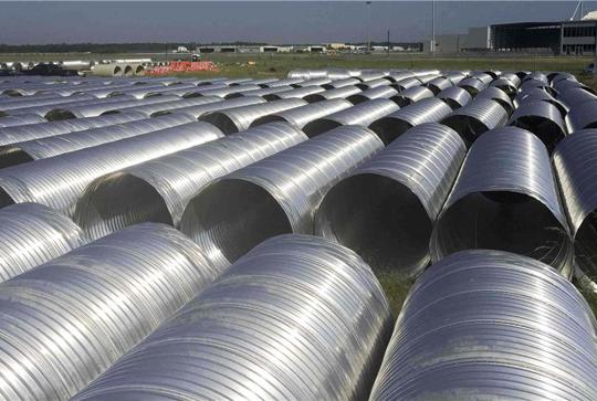 Large rows of shiny, cylindrical metal pipes arranged outdoors with a building in the background.