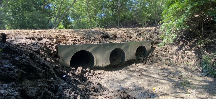 Three large concrete drainage pipes are installed in a muddy area surrounded by trees and vegetation.