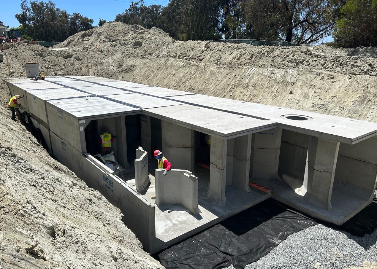 Workers assemble large concrete stormwater structures in a trench at a construction site under clear daylight.