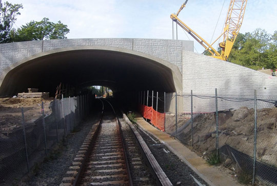A construction site with a newly built concrete railway bridge, surrounded by fencing and a crane in the background.
