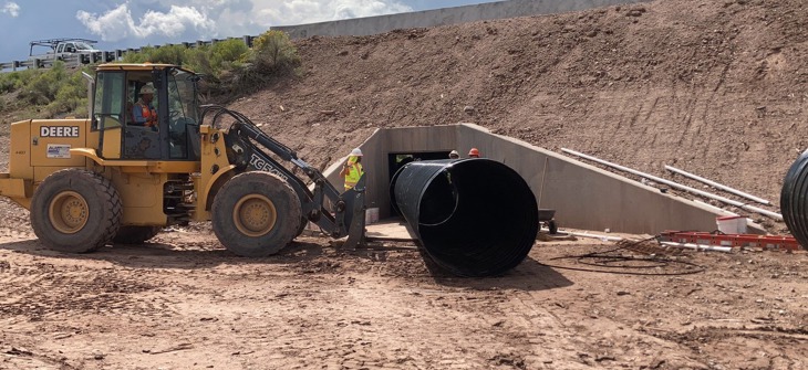 A front loader moves a large black pipe near a culvert under construction at a dirt worksite with several workers.