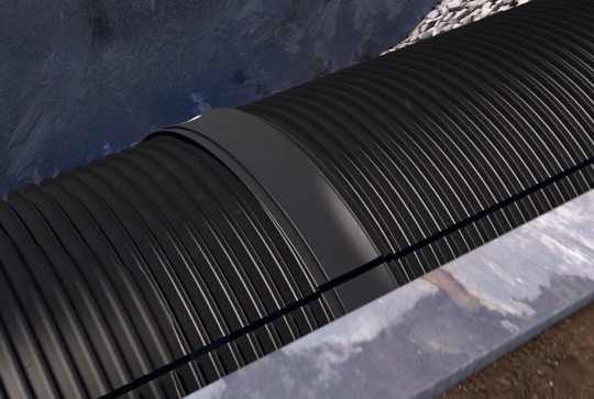 A black corrugated plastic pipe with a connector is installed in a trench lined with gravel and soil.