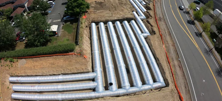 A construction site with large metal pipes laid out in parallel rows next to a road and nearby buildings.