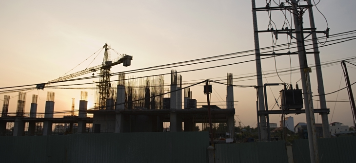 Construction site at sunset with cranes, unfinished building framework, and power lines silhouetted against the sky.