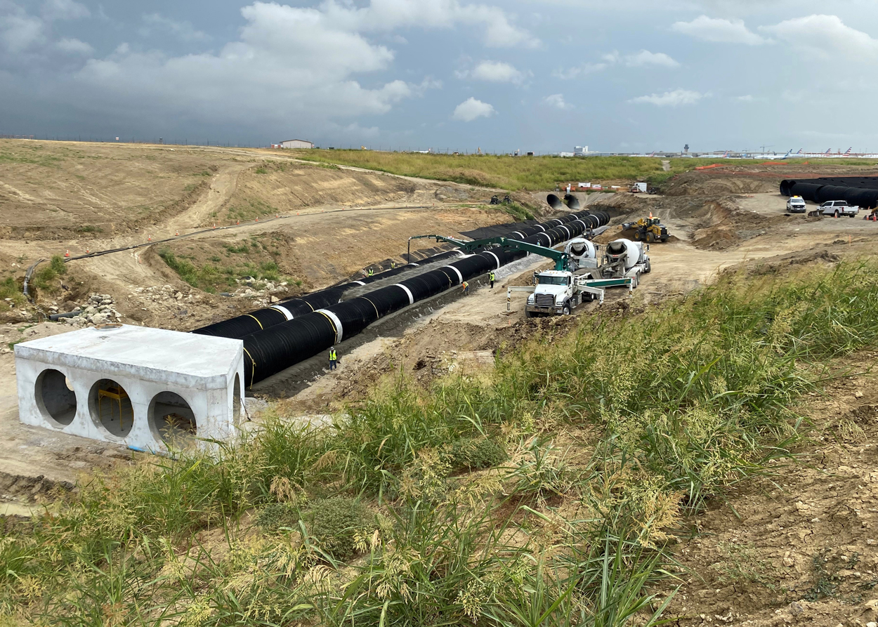 Large pipes and construction vehicles at a worksite in a cleared, grassy area under a cloudy sky.