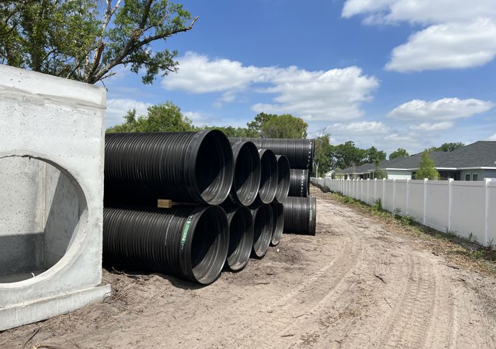 Large black plastic pipes are stacked beside a concrete structure at a construction site near a white fence and houses.