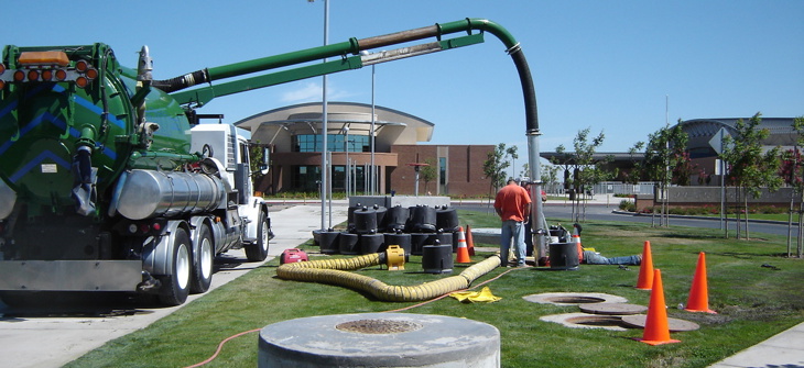 Worker operates vacuum truck to clean or service a manhole on a landscaped street, with safety cones set up nearby.