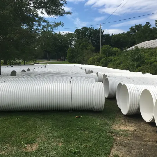 Large white corrugated plastic pipes are stacked in rows on grass near trees and power lines outdoors.