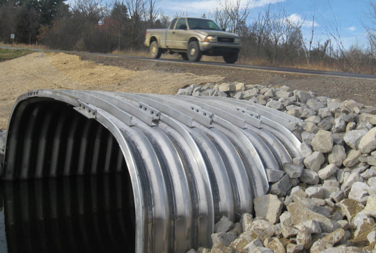 A truck drives over an arched metal culvert surrounded by rocks, allowing a stream to pass underneath a rural road.