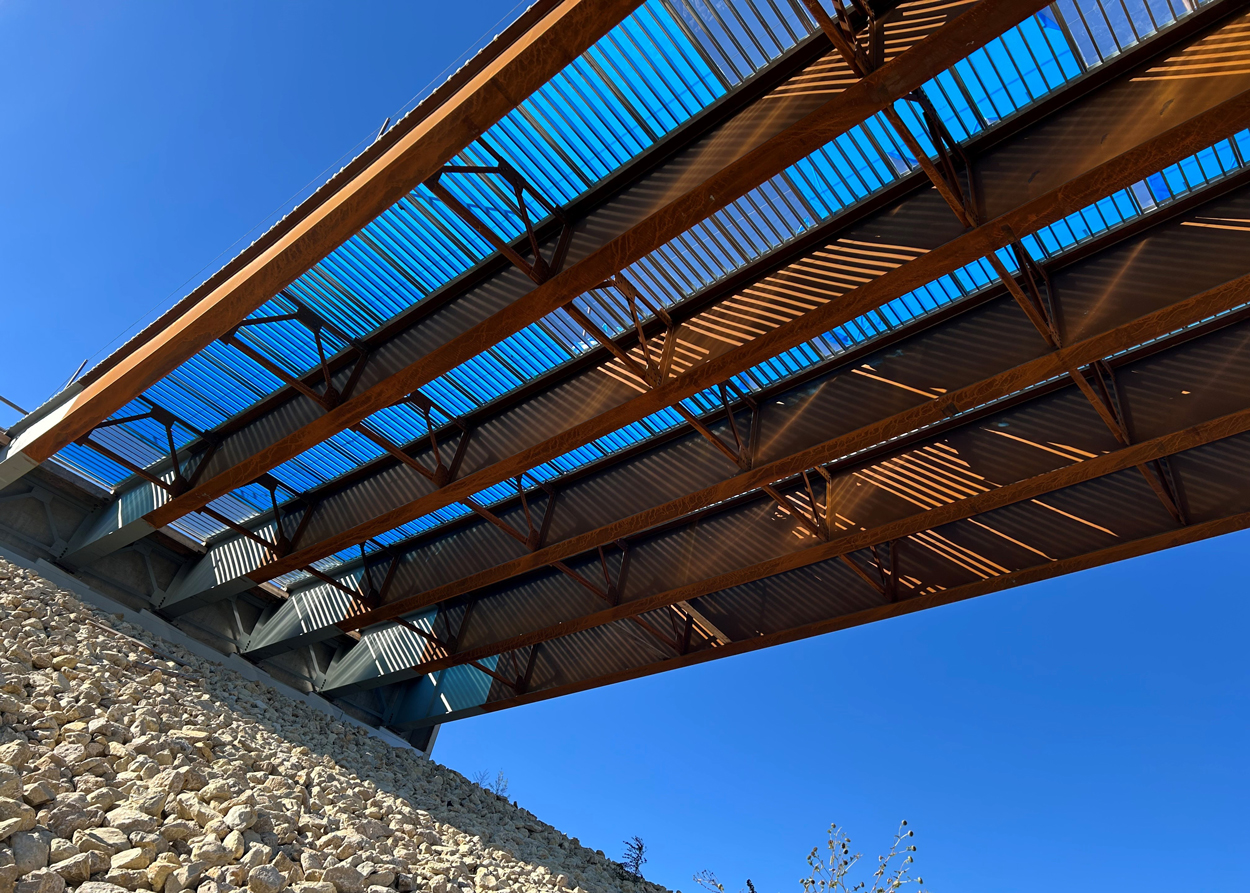 View from below a modern steel bridge with sunlight casting shadows, above a rocky embankment against a clear blue sky.