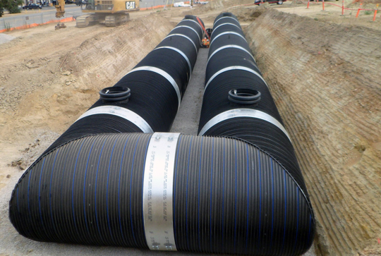 Large black plastic drain pipes installed in a trench at a construction site with soil banks on either side.