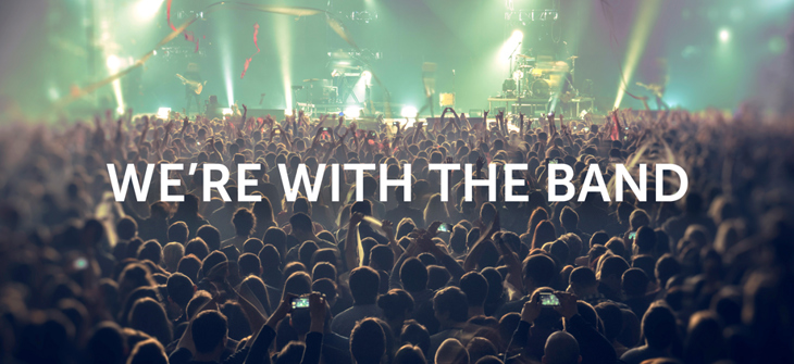 Crowd at a concert facing a brightly lit stage, with the text “WE’RE WITH THE BAND” overlaid in the center.