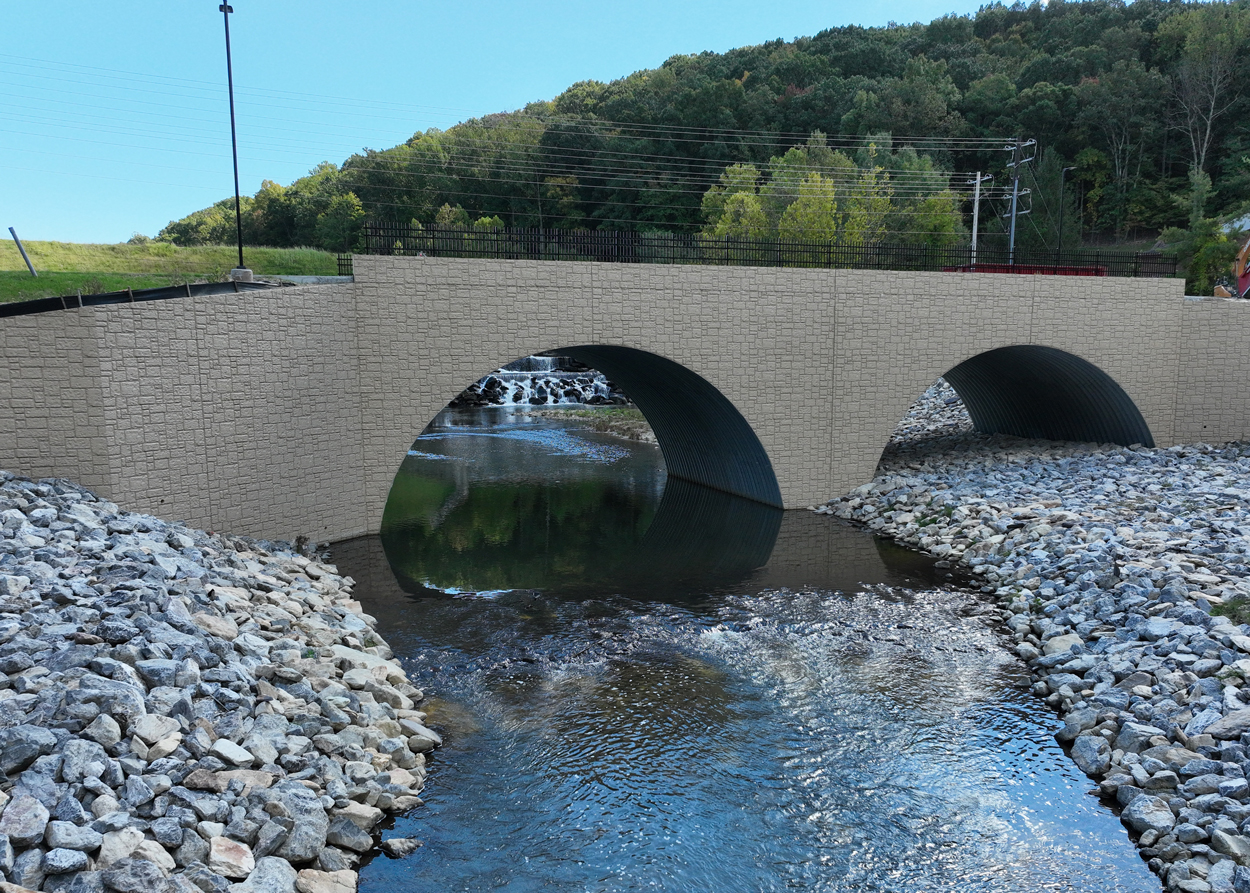 Stone-arch bridge with two arches spans a rocky stream, with trees and power lines in the background.