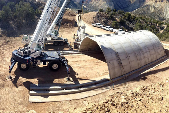 A crane positions a large concrete structure on a construction site surrounded by hills and trees.