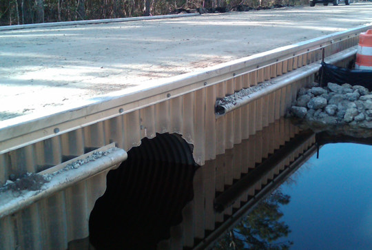 A small bridge with corrugated metal culvert and water underneath, bordered by orange traffic barrels and rocky shoreline.