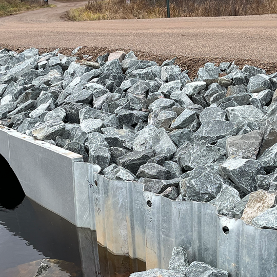 Culvert under a gravel road with large rocks lining the embankment, leading to water below.